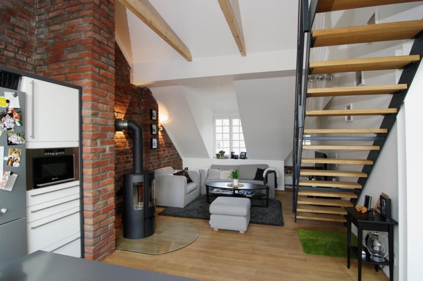 Das Prinzip Dachwohnung. Dachgeschossumbau im Denkmal., Düsseldorf-Oberrath — Wohnbereich mit freistehendem Kaminofen vor Ziegelwand und weißer Dachschräge