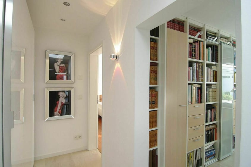 Umbau eines Bungalows, Duisburg-Rahm — Flur mit gerahmten Porträtfotos an der Wand, dahinter ein raumhohes Bücherregal